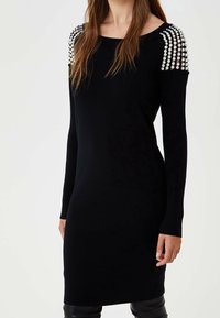 Robe en maille noire à manches longues, ornée d'accents de perles sur les épaules. Design simple et ajusté, texture lisse, coupe longueur genou.