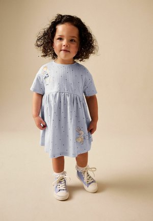 Toddler with curly hair wearing a light blue dress with polka dots and animal patches, paired with white socks and blue sneakers.