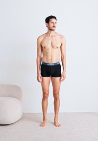 Fekete boxernadrág Lacoste logós derékrésszel. A modell mezítláb áll, semleges háttérrel és minimális bútorzattal.