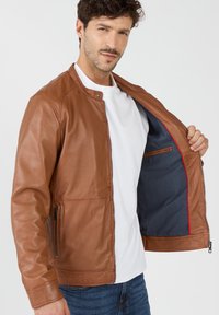 Braune Lederjacke mit Reißverschluss auf der Vorderseite, Stehkragen und zwei seitlichen Taschen. Innenfutter aus marineblauem Stoff mit roten Akzenten.