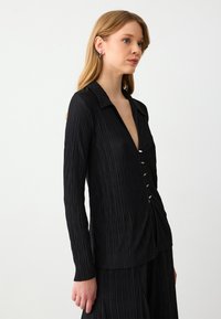 Zwarte geplooide blouse met lange mouwen en een kraag, voorzien van een v-halsopening en zilveren knopen. Gladde textuur, aansluitende silhouet.