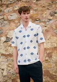 Young man wearing a white short-sleeve shirt with blue diamond patterns and dark pants, standing against a textured stone wall.