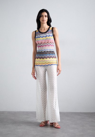 Stickad linne med flerfärgad zigzag-mönster; parat med genomskinliga vita vida byxor. Bärs med platta sandaler med rosa och orange detaljer.