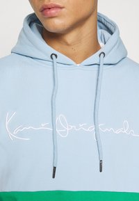 Felpa blu chiaro con cappuccio e lacci, con il logo ricamato "Kari Originals" in bianco; presenta un pannello inferiore verde e lacci con punte argentate.