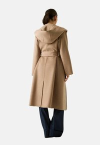 Cappotto di lana beige con cappuccio, vita con cintura e spacco laterale. Presenta maniche lunghe e una texture liscia. Indossato sopra pantaloni scuri a gamba larga.