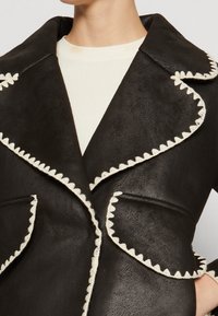 Femme portant un manteau noir texturé avec de grands revers et des coutures en zigzag blanches le long des bords, sur un haut blanc à côtes.