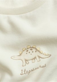 Ljusbeige tyg med ett tryckt stegosaurus i mjuk brunt med gula taggar, åtföljt av texten "Stegosaurus" i ett enkelt typsnitt.