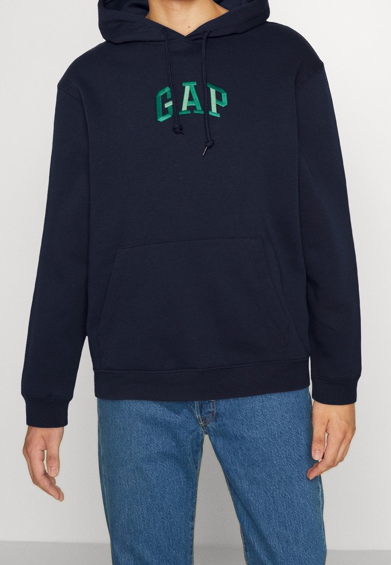 Personne portant un sweat à capuche bleu marine avec le logo vert "GAP" et un jean bleu, debout devant un fond clair uni.