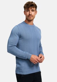 Lys blå strikket genser med et teksturert chevronmønster, rund halslinning og ribbede mansjetter og hem. Brukt med mørke bukser.