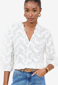 Weißes Bluse mit floralem Stickerei, langen Puffärmeln und V-Ausschnitt. Kombiniert mit blauen Jeans und einem beigen Gürtel. Goldfarbener Armreif als Akzent.