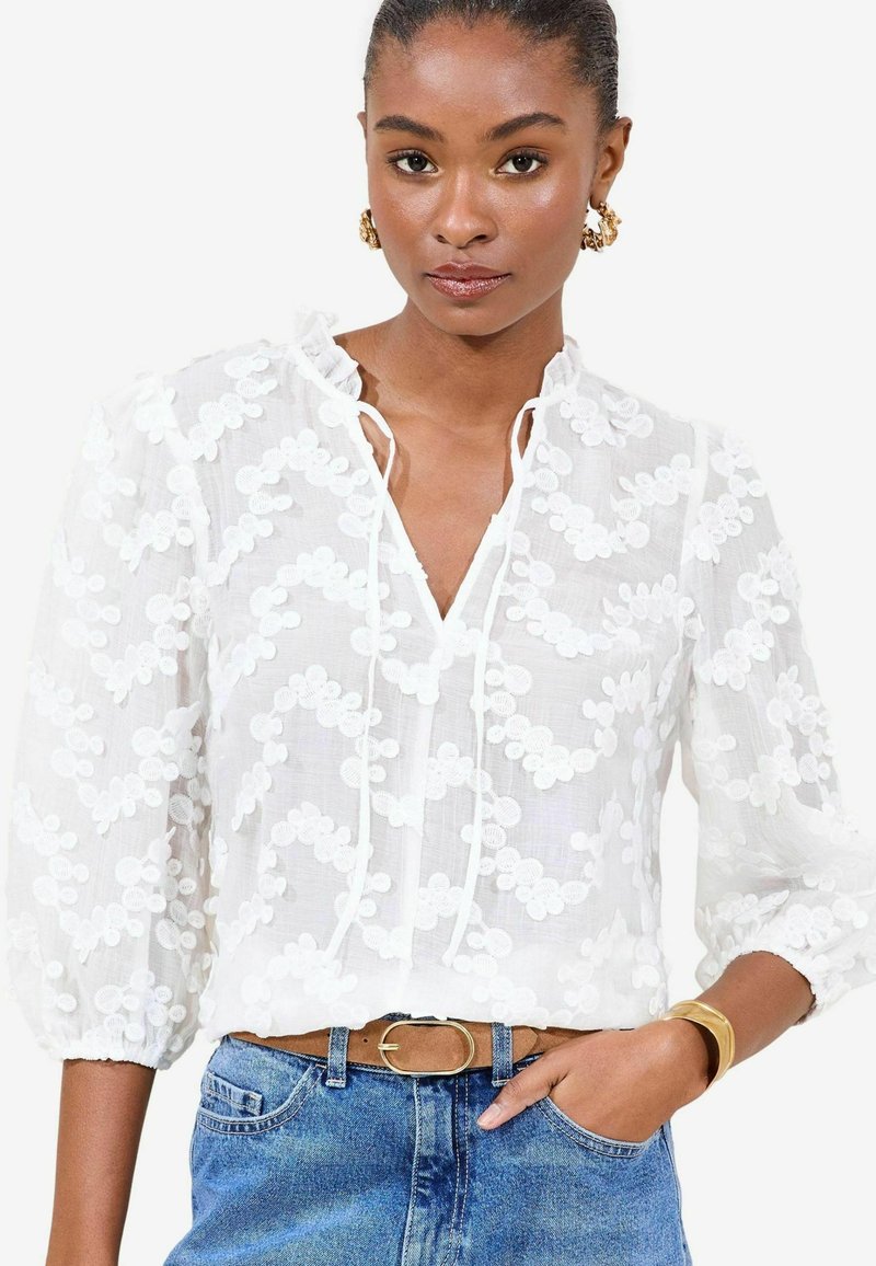 Weißes Bluse mit floralem Stickerei, langen Puffärmeln und V-Ausschnitt. Kombiniert mit blauen Jeans und einem beigen Gürtel. Goldfarbener Armreif als Akzent.