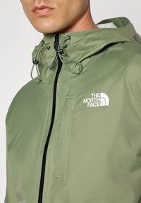 Light green waterproof jacket with a textured finish, black zipper, adjustable hood with drawstring, and a white logo on the chest.