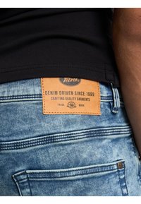 Jeans de mezclilla de color azul claro con una superficie texturizada, que presentan una etiqueta de cuero marrón etiquetada como "Tetroi®" y texto sobre la artesanía en denim.