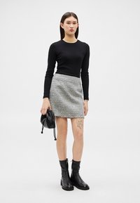 Γυναίκα με μαύρο μπλουζάκι μακρυμάνικο, γκρι μίνι φούστα από τουίντ, μαύρες μπότες, κρατώντας μαύρο τσαντάκι, στέκεται μπροστά σε απλό φωτεινό φόντο.