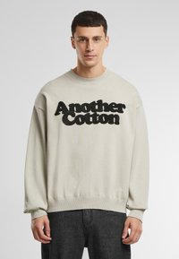 Fény szürke pamut pulóver, amelyen a mellrészen fekete textúrált "Another Cotton" logó található, valamint bordás mandzsetták és szegély.