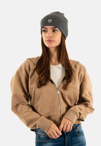 Jonge vrouw met een grijze beanie, een beige hoodie met ritssluiting over een wit T-shirt en een spijkerbroek, staand tegen een effen achtergrond.