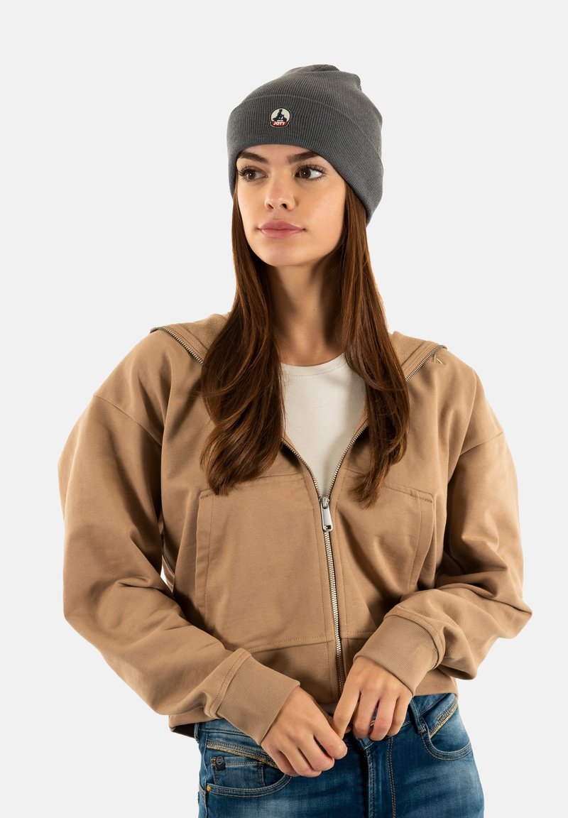 Jonge vrouw met een grijze beanie, een beige hoodie met ritssluiting over een wit T-shirt en een spijkerbroek, staand tegen een effen achtergrond.