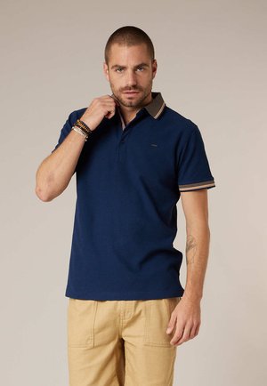 Homme ajustant son col, portant un polo bleu marine avec bordure rayée et pantalon beige, debout devant un fond uni.