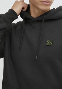 Uomo che indossa un hoodie nero con il patch verde "BLEND", che regola il cappuccio con una mano vicino al collo.