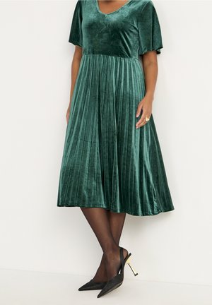 Kobieta ubrana w zieloną aksamitną plisowaną sukienkę midi z krótkimi rękawami, czarne przezroczyste rajstopy i czarne, spiczaste szpilki na złotych obcasach.