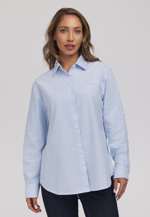 Kobieta o jasnobrązowych włosach, ubrana w jasnoniebieską koszulę z długim rękawem i ciemne spodnie na jednolitym tle.