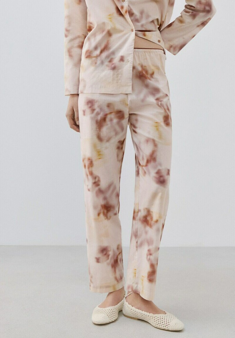 Femeie îmbrăcată într-un costum tie-dye bej și maro, cu pantaloni asortați și pantofi albi țesuti, stând în fața unui fundal simplu și deschis la culoare.