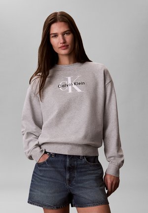 Ung kvinne med langt brunt hår iført en grå Calvin Klein genser og mørke denimbukser står mot en lys grå bakgrunn.
