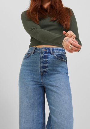 Persoon met lang rood haar, gekleed in een groen shirt met lange mouwen en een hoge taille blauwe spijkerbroek, handen samengevouwen voor een effen achtergrond.