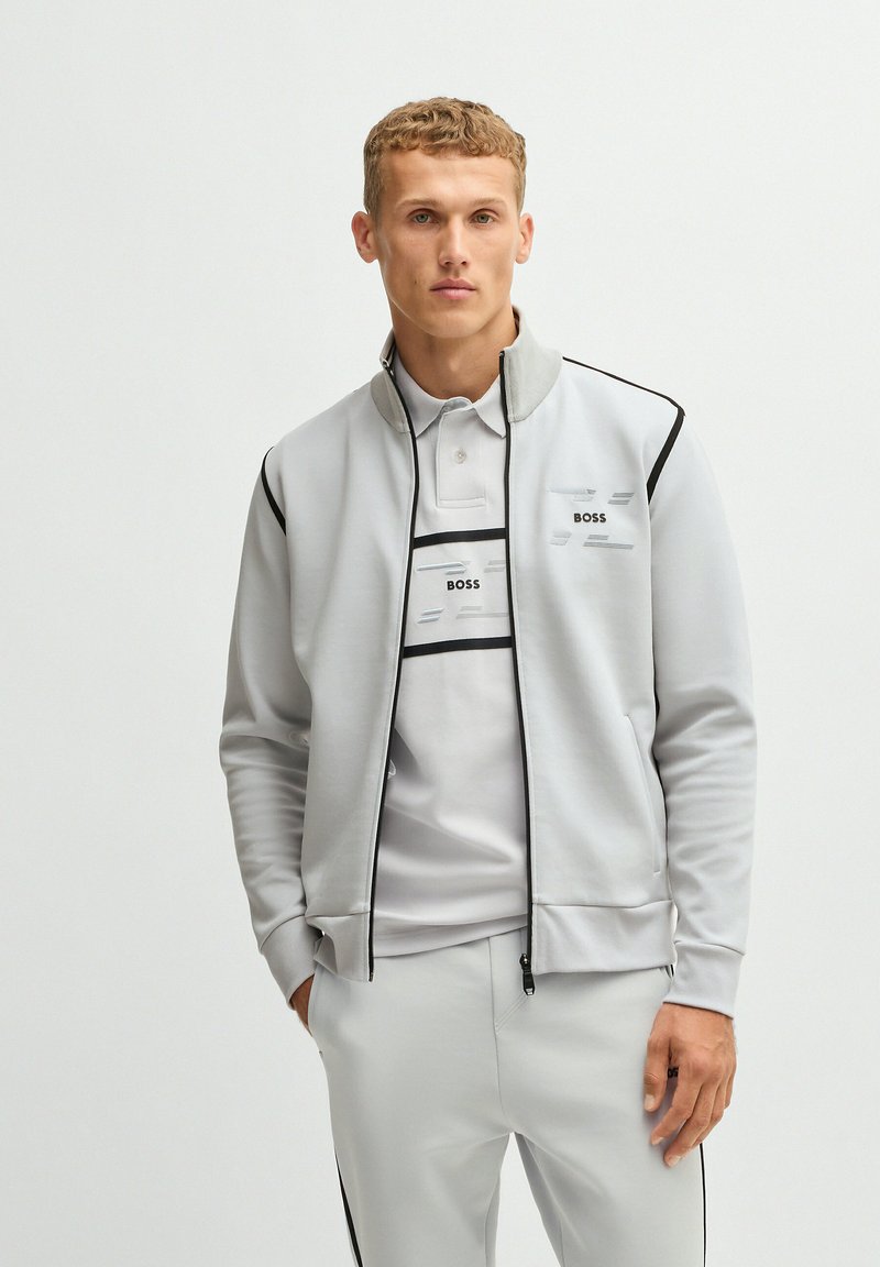 Veste zippée gris clair avec des accents noirs, poches avant et logo brodé. Associée à un polo assorti et un pantalon.