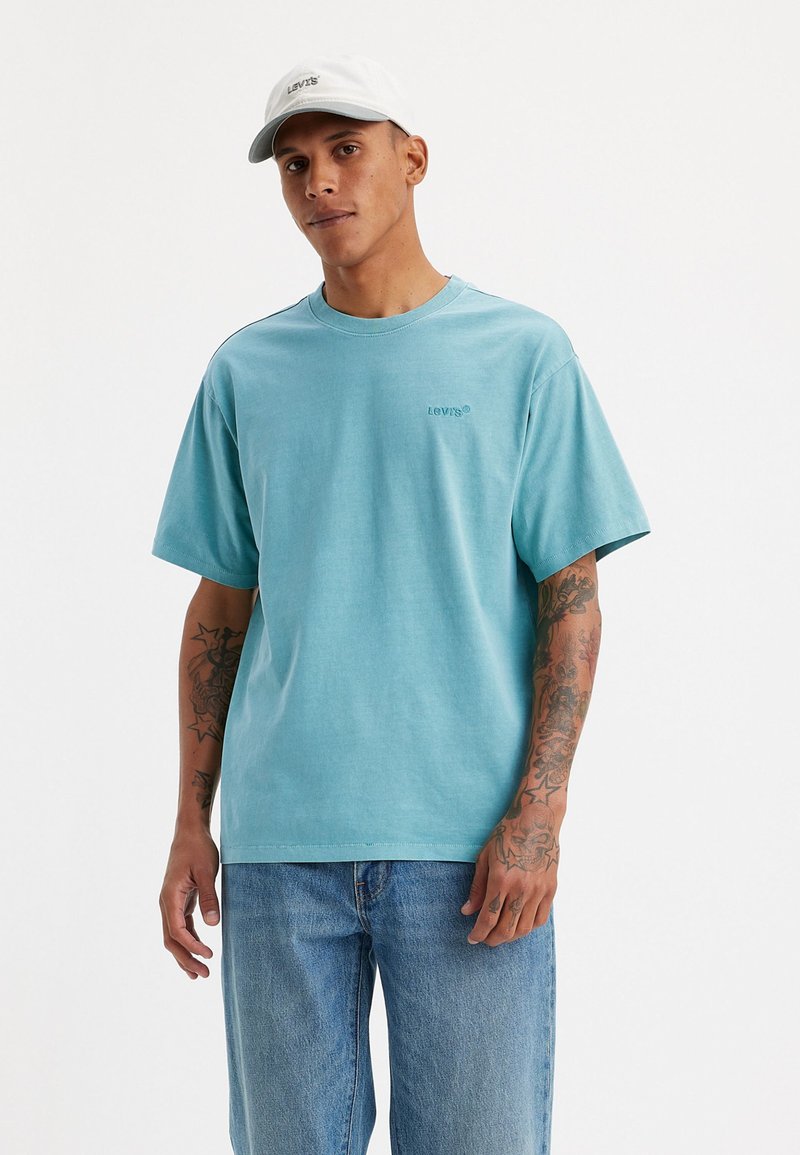 Tricou din bumbac turcoaz cu guler rotund și mâneci scurte, având un mic logo Levi's pe partea stângă. Poartă cu blugi albaștri.