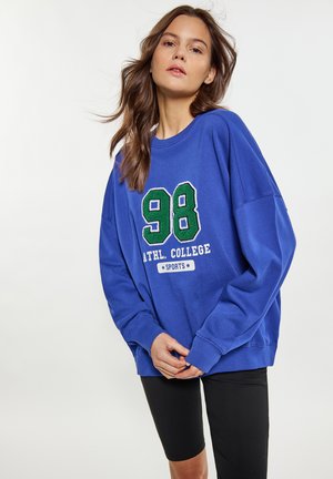 Kék kapucnis pulóver zöld és fehér hímzett szöveggel, amely az "98 ATHL. COLLEGE SPORTS." feliratot tartalmazza. Széles ujjakkal és laza szabással rendelkezik. Fekete sorttal párosítva.