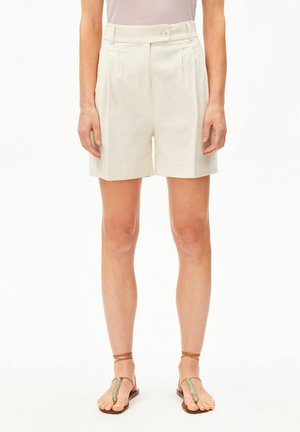 Personne portant un short crème taille haute plissé, un haut clair et des sandales marron avec brides à la cheville et perles décoratives.