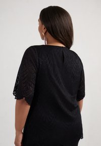 Top in pizzo nero con un motivo decorativo, maniche corte a scallop e dettaglio a occhiello sul retro. La texture sembra essere intrecciata in modo intricato.