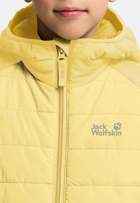 Chaqueta de plumas amarilla con cierre de cremallera, capucha y un sutil patrón acolchado. Logo impreso en gris en la parte frontal. Material suave y ligero.