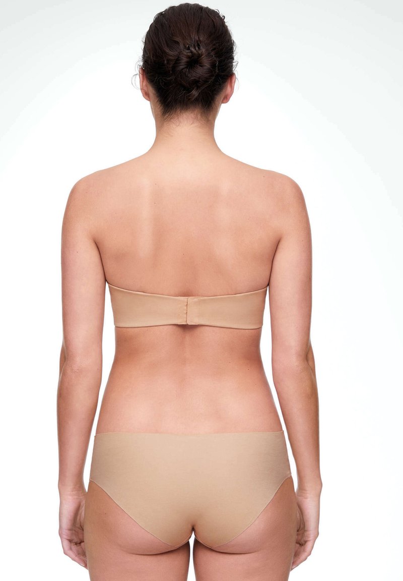 Femme aux cheveux foncés en chignon portant un soutien-gorge sans bretelles beige et des sous-vêtements assortis, vue de dos sur fond blanc.