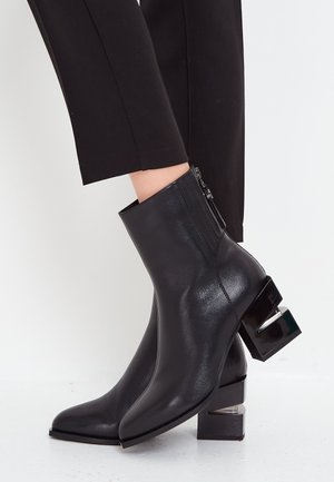 Schwarze Knöchelstiefel mit glänzender Lederoberfläche, quadratischer Zehenform, Reißverschluss und einem auffälligen quadratischen Blockabsatz mit metallischen Akzenten.