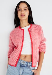 Austa rozā toņu adīta kardigāna uzvalks ar ribotu tekstūru, ar apaļu apkakli un pogu aizdari, valkāts virs baltā krekla.