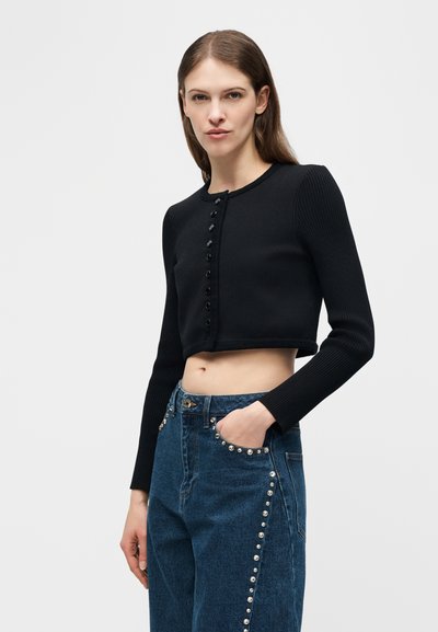 Γυναίκα που φοράει μαύρη κοντή ζακέτα με κουμπιά και ψηλόμεσο μπλε τζιν με ασημένιες τρουκς κατά μήκος των τσεπών και των πλευρών, στέκεται με το χέρι στην τσέπη.