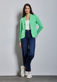 Lichtgroene blazer met twee zakken, gedragen over een witte top, in combinatie met wijde donkerblauwe jeans en witte sneakers. Eenvoudig ontwerp en pasvorm.