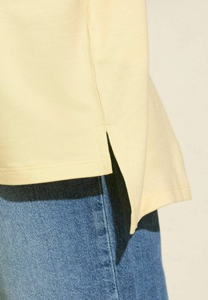 Světle žluté bavlněné tričko s bočním rozparkem a krátkými rukávy, v kombinaci s modrými dž�ínami s hladkou texturou a kontrastním prošíváním.