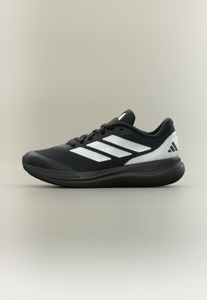 Chaussure de running Adidas noire et blanche avec trois bandes blanches et semelle rembourrée sur fond gris clair uni.