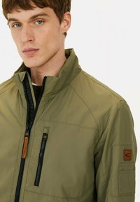 Olivegrüne leichte Jacke mit Stehkragen, Reißverschlusstoß und Seitentaschen. Verfügt über schwarze Reißverschlussbeschläge und sandfarbene Lederakzente.