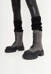 Sive suede čizme s vezicama i debelim crnim gumennim đonom. Imaju bočni zatvarač, teksturirane detalje i rebraste crne čarape.