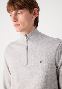 Pulover cu fermoar de culoare gri deschis, cu un design texturat, guler înalt și un logo mic brodat pe pieptul stâng. Detaliu cu fermoar argintiu.