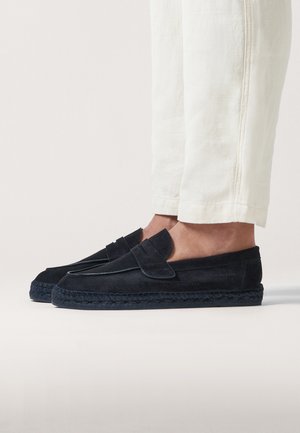 Chaussures slip-on en suède marine avec une semelle en jute tressé. Présente un rabat décoratif et des coutures minimales. Portées avec un pantalon couleur crème.