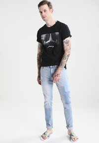 Camiseta negra con un gráfico de manos en oración, combinada con unos pantalones vaqueros claros desgastados y sandalias de deslizar con estampados. Presenta tatuajes visibles.