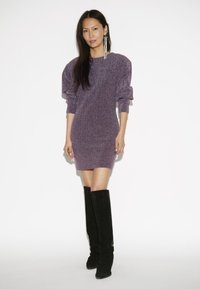 Vrouw in een paarse textuur lange mouwen mini-jurk en zwarte knielaarzen, staand tegen een witte achtergrond.