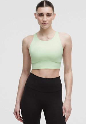 Mujer con el cabello oscuro recogido lleva un top corto sin mangas verde claro y leggings negros de cintura alta, de pie frente a un fondo liso.