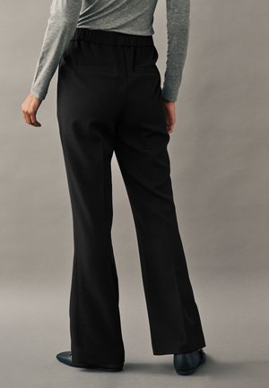 Persona che indossa pantaloni neri a gamba larga con vita elasticizzata, maglia grigia a maniche lunghe e scarpe nere, in piedi su uno sfondo semplice.