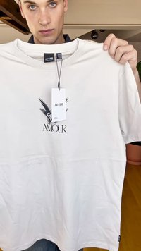 Weißes Baumwoll-T-Shirt mit lockerer Passform, das auf der Vorderseite mit einem schwarzen "AMOUR"-Grafik und einem Engel-Design versehen ist. Das Etikett ist am Kragen sichtbar.